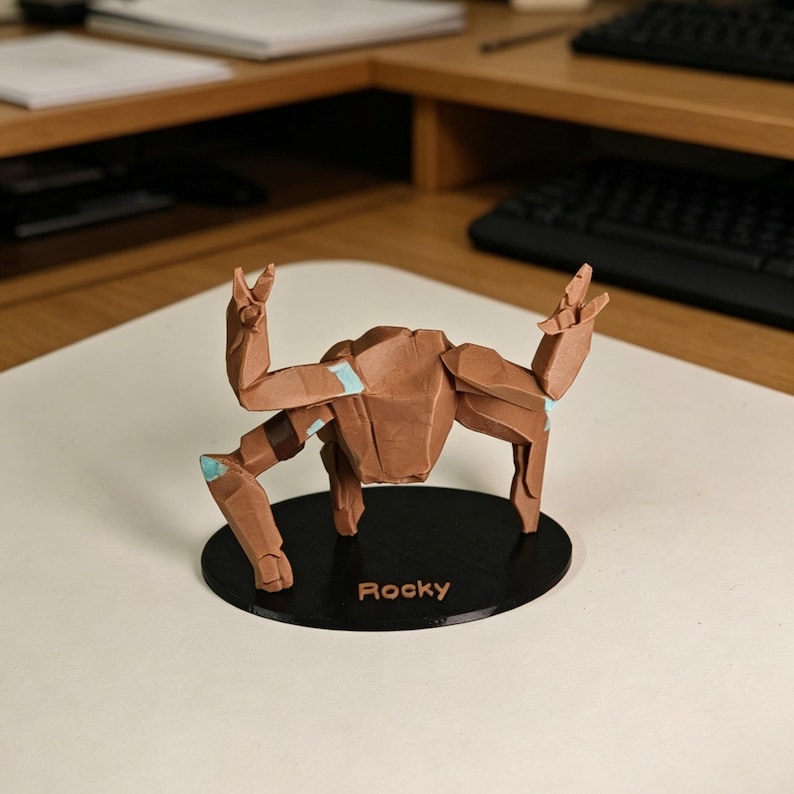 May include: A brown origami crab sculpture with light blue accents, mounted on a black circular base. The base is labeled "Rocky" in white text. The crab is posed with its claws raised.