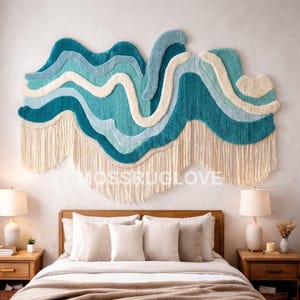 May include: A large, textured wall hanging featuring layered, wavy bands of teal, light blue, and cream yarn. The piece has a tufted, plush appearance with long, cream-coloured fringe hanging from the bottom edge. The text MUSSRUGLOVE is visible.