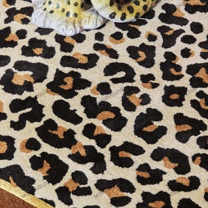 May include: A beige rug with a leopard print pattern in black and brown. The rug has a gold border and a decorative leopard statue is partially visible on top of the rug.