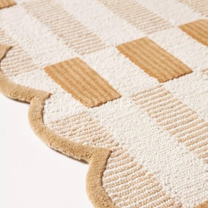 Puede incluir: Una alfombra beige y blanca estampada con borde festoneado. La alfombra presenta un dise&ntilde;o de tablero de ajedrez con bloques rectangulares beige y blancos. El borde de la alfombra es de color beige liso.