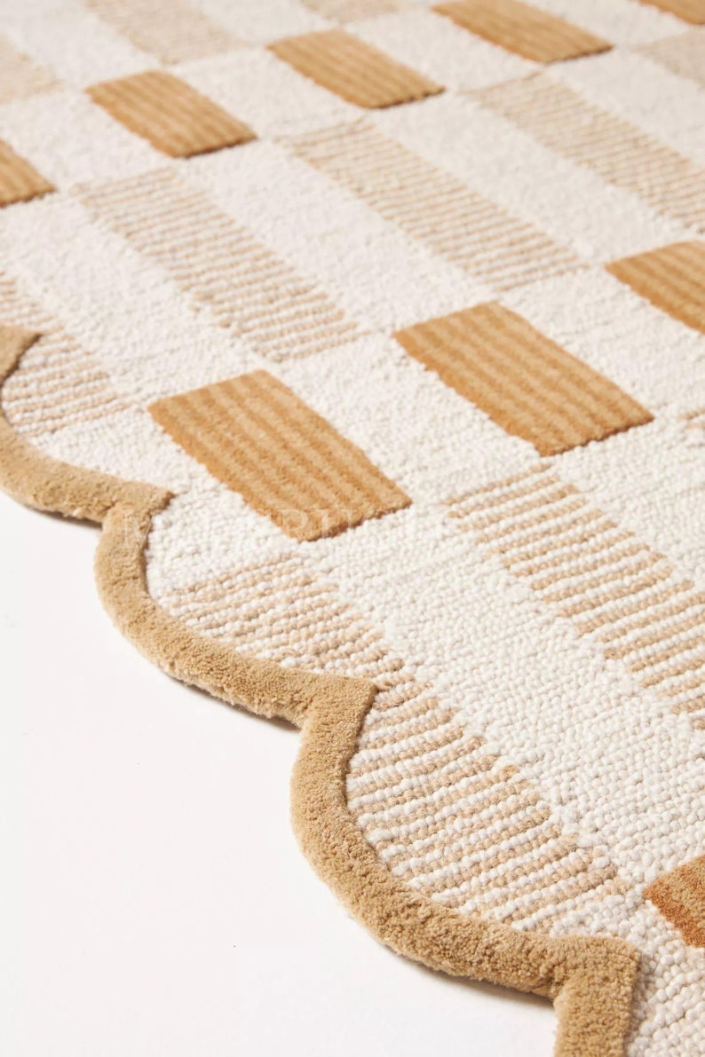 Puede incluir: Una alfombra beige y blanca estampada con borde festoneado. La alfombra presenta un dise&ntilde;o de tablero de ajedrez con bloques rectangulares beige y blancos. El borde de la alfombra es de color beige liso.