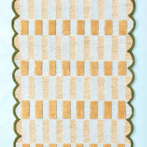 Puede incluir: Una alfombra rectangular con un patr&oacute;n repetitivo de rayas verticales blancas y doradas, intercaladas con bloques rectangulares dorados. La alfombra tiene un borde verde festoneado, creando un borde decorativo. La alfombra est&aacute; sobre una superficie azul claro.