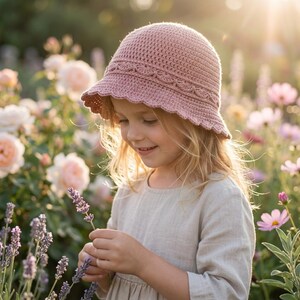 Op de afbeelding: Een lichtroze gehaakte hoed met een geschulpte rand en decoratieve band. De hoed wordt gedragen door een persoon in een bloemenveld. De hoed heeft een zachte, gedempte roze kleur.