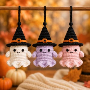 May include: Three crocheted ghost ornaments hanging from a wooden rod. Each ghost features a black witch hat with an orange band and buckle, black bead eyes, and a stitched smile. The ghosts are colored white, light purple, and light pink.