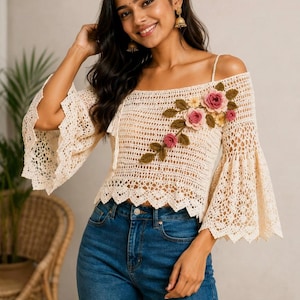 Puede incluir: Top de crochet color crema con hombros descubiertos y mangas acampanadas. El top presenta una aplicación floral con rosas rosas y amarillas y hojas verdes. La modelo lleva vaqueros azules. El top es de estilo bohemio.