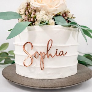 May include: A white iced cake decorated with a floral arrangement, featuring a cream rose and foliage. A copper-coloured cake topper spells "Sophia" in cursive. The cake is presented on a grey plate. The cake is approximately 15 cm in diameter.