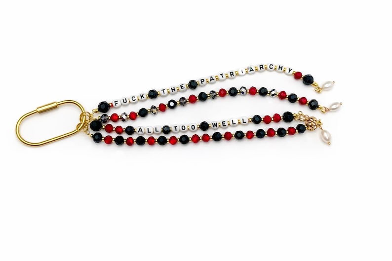 May include: Gold-toned keychain with three beaded strands. The strands feature black, red, and white beads spelling out phrases. Each strand is adorned with a small pearl charm. The text reads "FUCK THE PATRIARCHY" and "ALL TOO WELL".