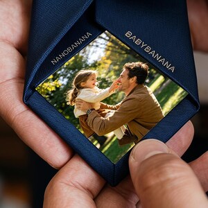 May include: A navy blue necktie featuring a hidden interior panel with a printed photograph of an adult and a child outdoors. The fabric edges of the inner flap are inscribed with the text NANOBANANA and BABYBANANA in a clean, sans-serif font.