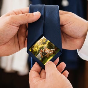 Puede incluir: Una corbata azul marino lisa con una inserción fotográfica rectangular oculta en la parte inferior de la pala ancha. La fotografía muestra a dos personas sentadas al aire libre en un jardín soleado con árboles y follaje de fondo.