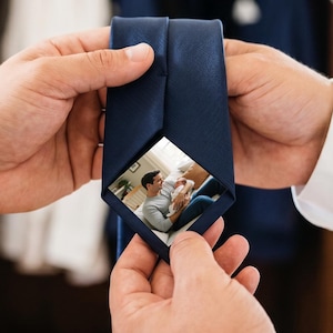 Puede incluir: Una corbata azul marino lisa con una inserción fotográfica rectangular oculta en el forro interior. La fotografía muestra a una persona sosteniendo a un bebé mientras está sentada en un sofá. La corbata es de tela lisa y se mantiene abierta.