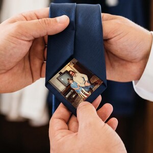 Puede incluir: Una corbata de color azul marino sólido que presenta una pequeña fotografía rectangular impresa en la punta frontal. La fotografía muestra a dos personas de pie en un interior, una con camisa azul claro y la otra con camisa blanca.