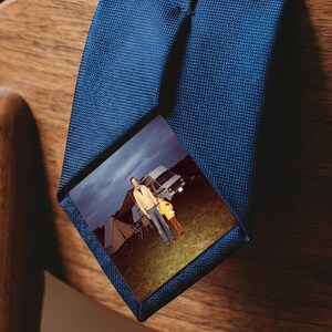 Puede incluir: Una corbata azul marino lisa con una impresión fotográfica rectangular en la pala. La fotografía muestra a una persona de pie en un campo de hierba junto a una tienda de campaña y un vehículo bajo un cielo oscuro y nublado. Tejido texturizado.