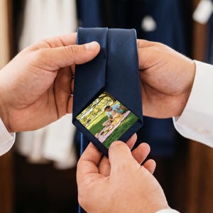 Puede incluir: Una corbata azul marino lisa con una foto rectangular oculta en la parte interior. La imagen muestra a una pareja haciendo un picnic en un césped verde con árboles de fondo. La corbata está confeccionada con un tejido liso y mate.
