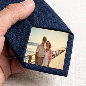May include: A dark blue necktie featuring a textured fabric finish and a rectangular photo insert on the bottom tip. The photo depicts a couple standing on a wooden pier by the ocean, with the man in a light shirt and the woman in a white dress.