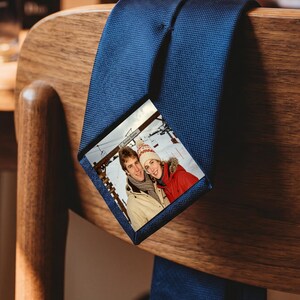 Puede incluir: Una corbata azul marino lisa con una inserción fotográfica rectangular en la parte delantera. La fotografía muestra a dos personas con ropa de invierno, incluyendo un gorro de punto y una chaqueta, en un entorno nevado con telesillas.