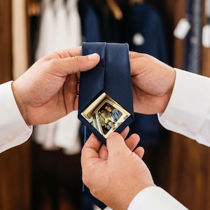 Puede incluir: Una corbata azul marino lisa que presenta una pequeña fotografía cuadrada con marco dorado incrustada en la tela cerca de la punta. La fotografía muestra una imagen clara de dos personas sentadas dentro de un vehículo, visible a través del marco.