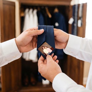 Puede incluir: Una corbata azul marino lisa con una fotografía rectangular oculta cosida en la parte inferior del extremo ancho. La fotografía muestra a dos personas de pie juntas, una con una camisa amarilla y la otra con una blusa de color rosa.