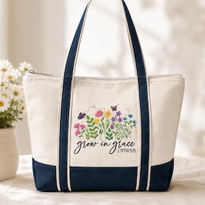 May include: A cream-colored canvas tote bag with navy blue trim and handles. The front features a colorful embroidered design of various wildflowers and two butterflies, with the black text "grow in grace" and "2 PETER 3:18" printed below.