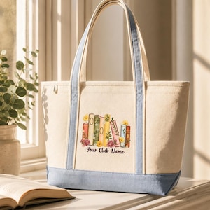 Op de afbeelding: Een beige canvas draagtas met lichtblauwe randen en hengsels. De voorzijde is voorzien van een afbeelding van een rij kleurrijke boeken versierd met bloemen en bladeren, met de tekst "Your Club Name" in een donker lettertype eronder.