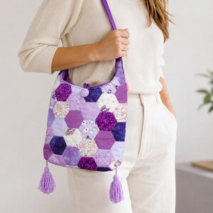 Peut inclure: Un sac fourre-tout en patchwork avec une bandoulière et des pompons violets. Le sac est composé de pièces de tissu hexagonales dans différentes nuances de violet et de motifs floraux.