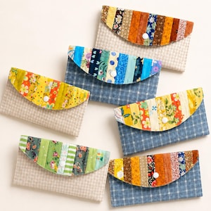May include: Five rectangular fabric wallets with rounded envelope-style flaps. Each flap features vertical strips of colorful patterned fabric. The wallet bodies are made of either beige or blue plaid fabric, each secured with a white snap button.