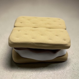 May include: A ceramic s'more with two tan graham cracker squares and a white marshmallow with a brown chocolate layer. The graham crackers have a textured surface with small dimples.