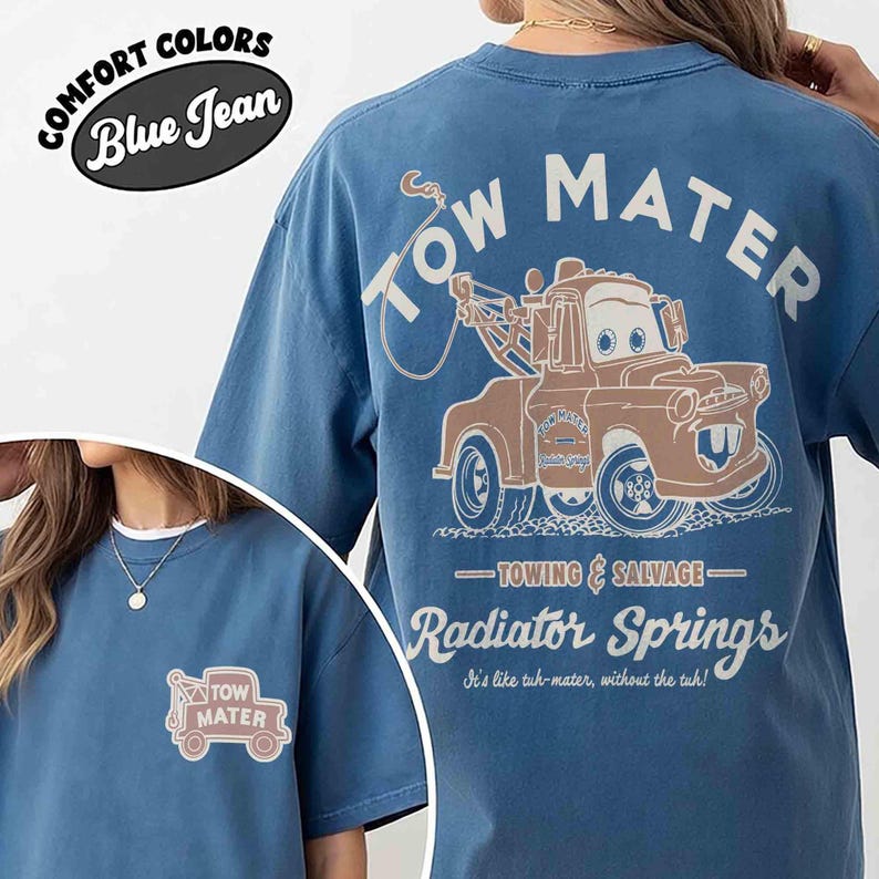 May include: A blue jean colored t-shirt featuring a graphic of the character Tow Mater on the back with the text Tow Mater, Towing & Salvage, Radiator Springs, and It's like tuh-mater, without the tuh. A smaller Tow Mater graphic is on the front.