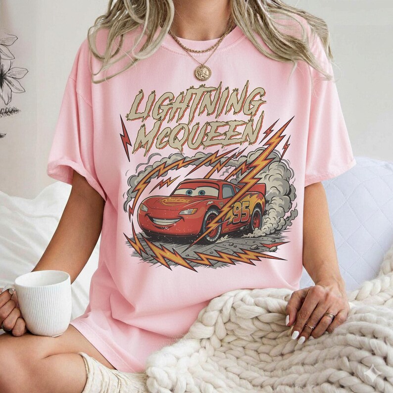 May include: Pink t-shirt featuring a graphic of Lightning McQueen from the Cars movie. The design includes the text "Lightning McQueen" above the car, with lightning bolts and smoke effects. The car is red with the number 95.