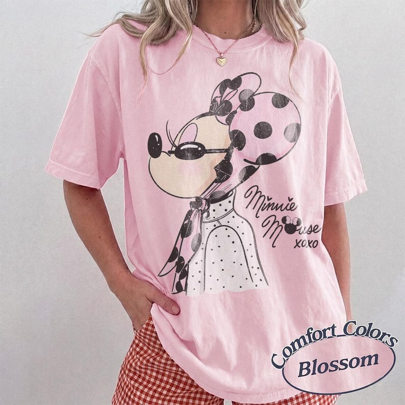 May include: A light pink t-shirt featuring a Minnie Mouse graphic with a black and white polka dot bow and scarf. The text "Minnie Mouse xoxo" is printed on the shirt. The shirt also has the text "Comfort Colors Blossom".