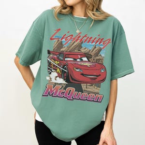 K&ouml;nnte beinhalten: Salbeigr&uuml;nes T-Shirt mit einem Aufdruck von Lightning McQueen aus dem Film Cars. Das Design zeigt den Text "Lightning McQueen" in Rot und Lila, vor einer W&uuml;stenlandschaft.