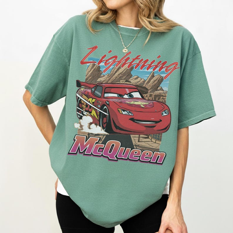 K&ouml;nnte beinhalten: Salbeigr&uuml;nes T-Shirt mit einem Aufdruck von Lightning McQueen aus dem Film Cars. Das Design zeigt den Text "Lightning McQueen" in Rot und Lila, vor einer W&uuml;stenlandschaft.