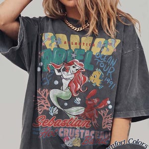 May include: Dark gray t-shirt featuring a colorful graphic of Ariel from The Little Mermaid, with the text "Ariel Ocean Tour 1991". The design includes characters like Flounder and Sebastian. The shirt is labeled "Comfort Colors Pepper".