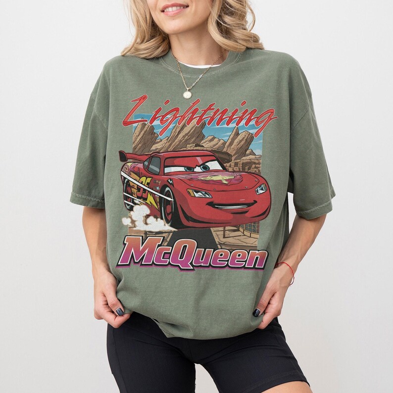 K&ouml;nnte beinhalten: Olivgr&uuml;nes T-Shirt mit einem Aufdruck von Lightning McQueen aus dem Film Cars. Das Design enth&auml;lt den Text "Lightning McQueen" in Rot und "McQueen" in Lila. Das T-Shirt hat einen Vintage-Look.
