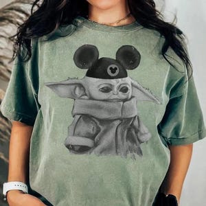 May include: A sage green short-sleeved t-shirt featuring a graphic print of a small, large-eared creature wearing a black cap with round ears and a small circular icon. The shirt has a distressed, washed-out texture and a crew neckline.
