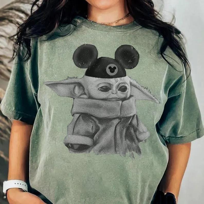 May include: A sage green short-sleeved t-shirt featuring a graphic print of a small, large-eared creature wearing a black cap with round ears and a small circular icon. The shirt has a distressed, washed-out texture and a crew neckline.