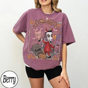 K&ouml;nnte beinhalten: Beerenfarbenes T-Shirt mit einem Halloween-Motiv. Das Design zeigt Zeichentrickfiguren, einen Grabstein mit dem Text "NAME 2026" und K&uuml;rbisse. Das Shirt hat eine lockere Passform und kurze &Auml;rmel.