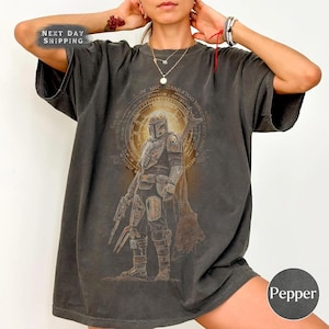 May include: A charcoal gray short-sleeve t-shirt featuring a graphic print of a helmeted warrior in armor standing in front of a circular golden emblem. The design includes intricate line work and a weathered, vintage-inspired aesthetic.
