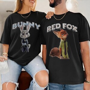 以下が含まれることがあります： ズートピアのキャラクターが描かれた黒いTシャツ。片方のシャツにはジュディ・ホップスと「BUNNY」の文字が、もう片方のシャツにはニック・ワイルドと「RED FOX」の文字が描かれています。