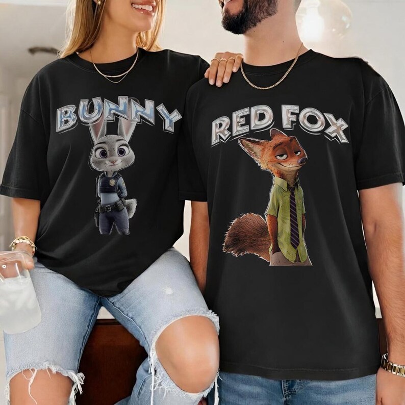 以下が含まれることがあります： ズートピアのキャラクターが描かれた黒いTシャツ。片方のシャツにはジュディ・ホップスと「BUNNY」の文字が、もう片方のシャツにはニック・ワイルドと「RED FOX」の文字が描かれています。