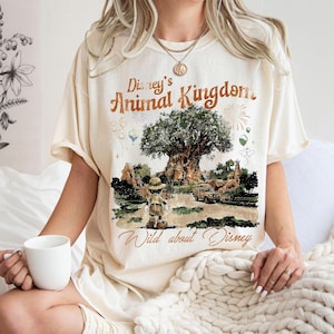 May include: Cream-colored t-shirt featuring a graphic of Disney's Animal Kingdom with the Tree of Life, Mickey Mouse, and the text "Disney's Animal Kingdom" and "Wild about Disney". The shirt has short sleeves and a relaxed fit.