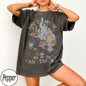 May include: Dark gray t-shirt featuring a colorful graphic of cartoon animals and the text "WHERE ANYONE CAN BE ANYTHING". The design includes a cityscape and various characters. The shirt has short sleeves and a relaxed fit.