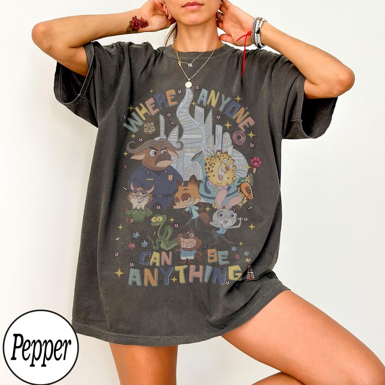 May include: Dark gray t-shirt featuring a colorful graphic of cartoon animals and the text "WHERE ANYONE CAN BE ANYTHING". The design includes a cityscape and various characters. The shirt has short sleeves and a relaxed fit.