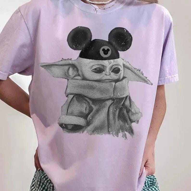 May include: A light purple t-shirt featuring a black and white graphic print of a small creature with large ears wearing a black cap with round ears and a small circular icon. The creature is depicted in a sketch style with a textured appearance.