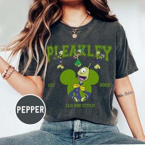 May include: Dark gray t-shirt featuring a green Pleakley graphic from Lilo and Stitch. The design includes the word "PLEAKLEY" above the character, with "EST 2002" and "LILO AND STITCH" below. The graphic is in a Mickey Mouse shape.