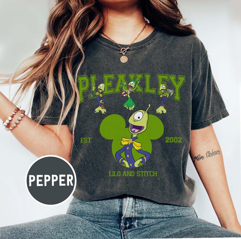 May include: Dark gray t-shirt featuring a green Pleakley graphic from Lilo and Stitch. The design includes the word "PLEAKLEY" above the character, with "EST 2002" and "LILO AND STITCH" below. The graphic is in a Mickey Mouse shape.