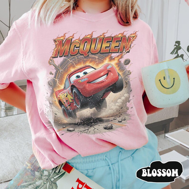Op de afbeelding: Een lichtroze t-shirt met een afbeelding van een rode raceauto met het nummer 95 en de tekst MCQUEEN in vetgedrukte letters. Het ontwerp bevat vlammen, gereedschap en een stoffig grondeffect. Een lichtblauwe mok met een gele smiley.