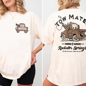 May include: An off-white t-shirt featuring a small graphic of a brown tow truck on the front left chest. The back displays a large brown tow truck graphic with the text Tow Mater, Towing & Salvage, Radiator Springs, and It's like tuh-mater, without the tuh.