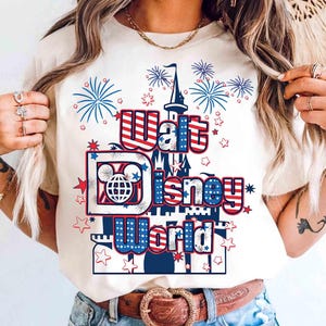 May include: Cream-colored t-shirt featuring the words "Walt Disney World" in red, white, and blue, with a castle graphic and fireworks. The design includes the iconic Mickey Mouse globe logo and star accents.