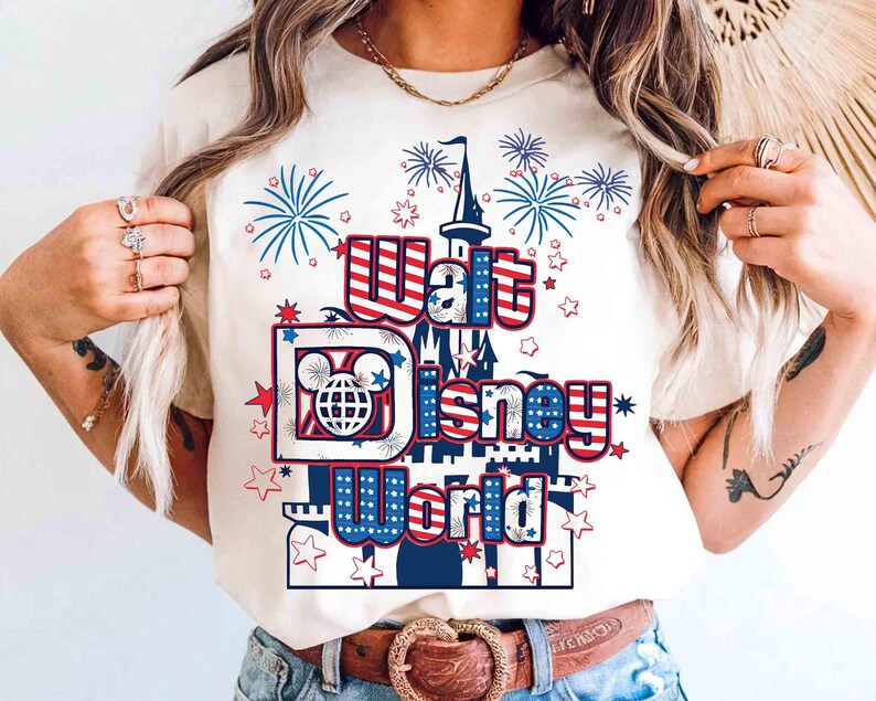 May include: Cream-colored t-shirt featuring the words "Walt Disney World" in red, white, and blue, with a castle graphic and fireworks. The design includes the iconic Mickey Mouse globe logo and star accents.