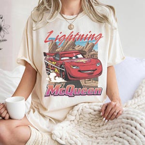 K&ouml;nnte beinhalten: Cremefarbenes T-Shirt mit einer Grafik von Lightning McQueen aus dem Film Cars. Das Design enth&auml;lt den Text "Lightning McQueen" in Rot und "McQueen" in Lila. Das Auto ist rot mit gelben und schwarzen Akzenten.